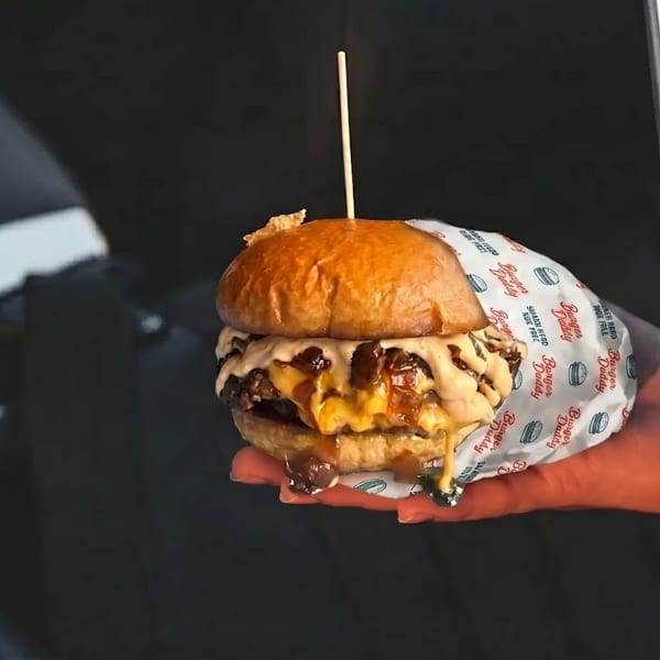A hand holds a Burger Daddy double cheeseburger with melted cheese and sauce, wrapped in branded paper.
